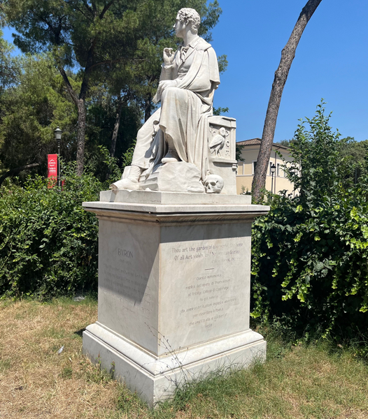 Estatua de mármol blanco de Lord Byron sentado en unas ruinas griegas, vestido con traje y capa, con una calavera y una lechuza en el pedestal donde está sentado. 