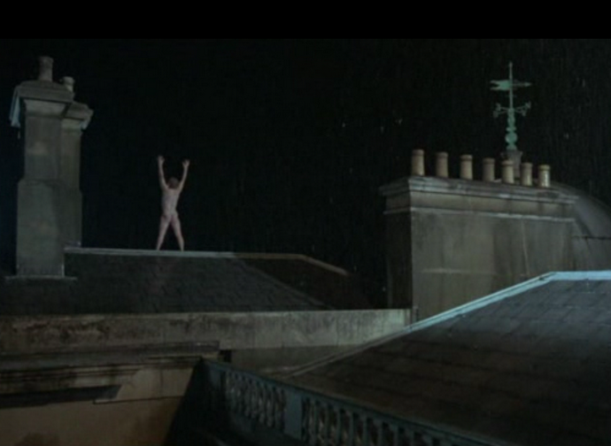 Fotograma con Julian Sands desnudo de espaldas con el cuerpo estirado y os brazos hacia arriba, invocando a una tormenta subido a un tejado