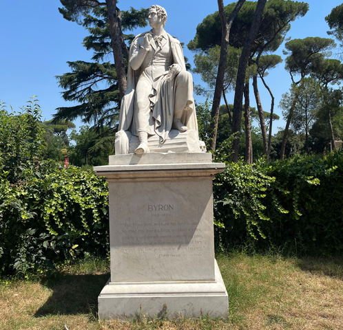 Estatua de mármol de Lord Byron sentado sobre un pedestal, en un jardín rodeado de árboles y vegetación. En el pedestal aparece la inscripción «BYRON 1788–1824» junto a un fragmento de una de sus obras, Childe Harold
