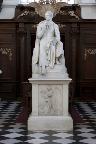 Estatua de mármol blanco de Lord Byron sentado, vestido con traje y capa, sosteniendo un libro enrollado. La escultura se encuentra en el interior de la biblioteca en madera frente a una vidriera decorativa.