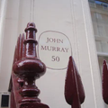 Detalle de la fachada de la editorial Murray, donde detrás de una verja roja, se muestra un circulo con John Murray 50 en su interior.