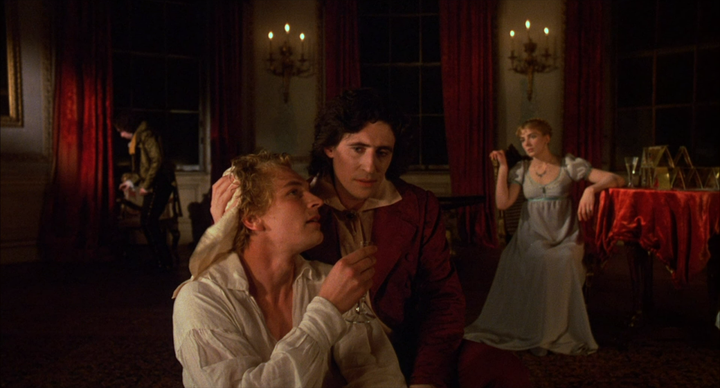 Imagen de los actores que encarnan a Byron y a Shelley que muestra uno de los ataques que sufría Shelley mientras Byron le atiende con un pañuelo secándole el sudor. Mary Shelley se encuentra al fondo de la estancia mirando a ninguna parte.