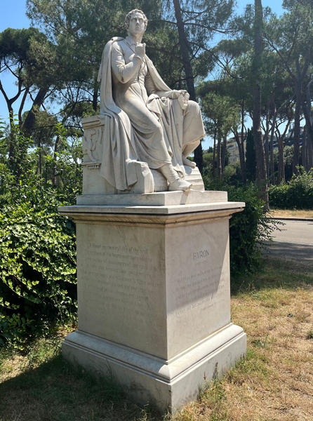 Estatua de mármol blanco de Lord Byron sentado en unas ruinas griegas, vestido con traje y capa, con un grifo y una lira en el pedestal donde está sentado